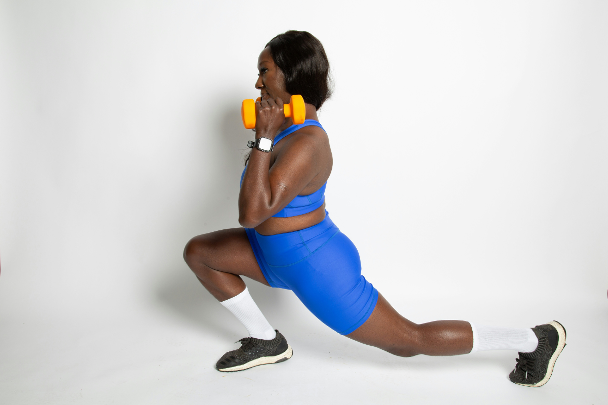 A woman of colour does a lunge. She is holding orange weights near her shoulders and is wearing a blue outfit with white socks and black shoes. We are seeing her from the side.