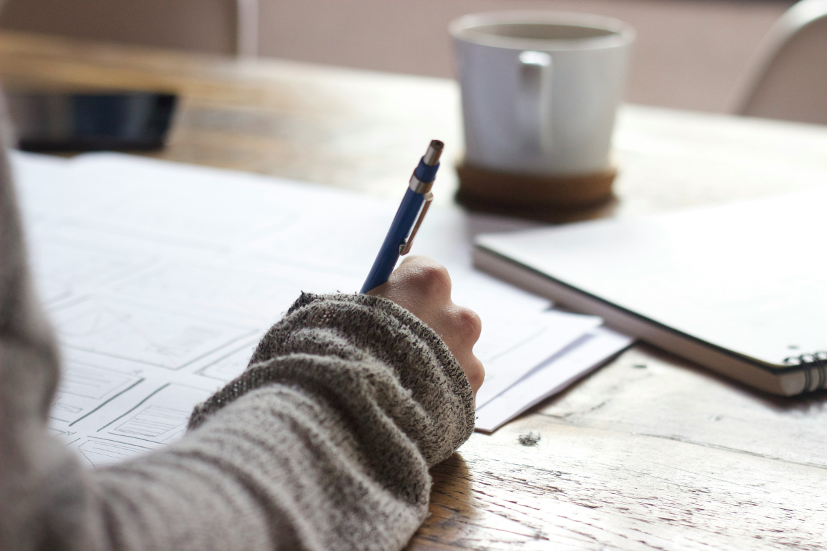 A light skinned hand writes on paper with a pen on a wooden desk. There is a coffee cup in the background.