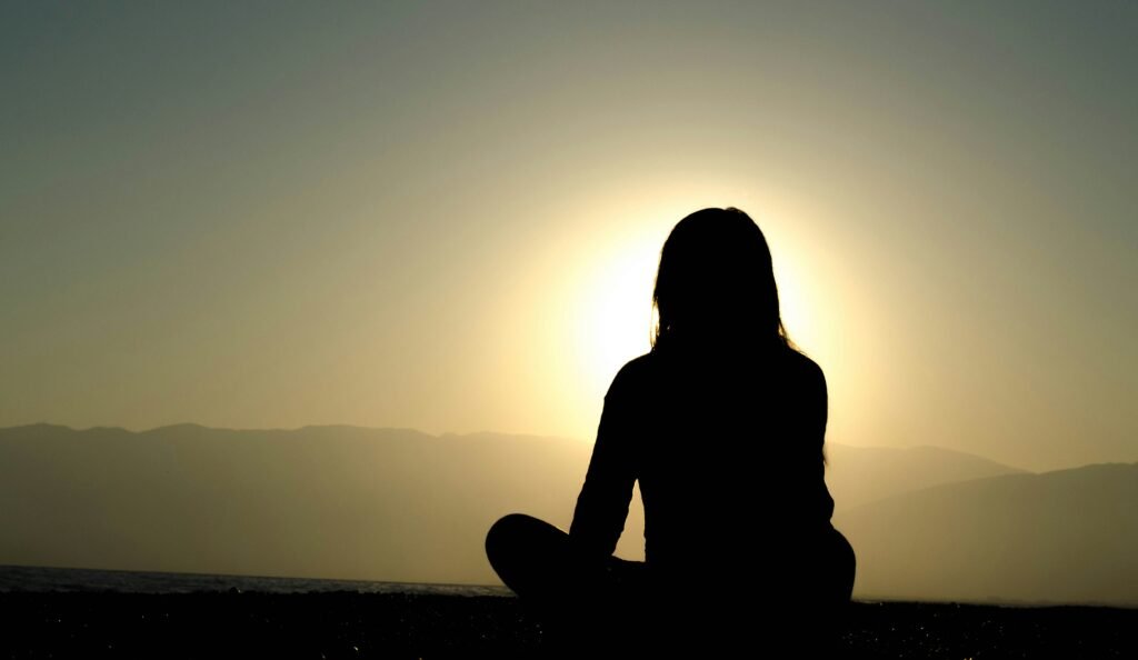 sharing from the scar not the wound image: photograph a person with long hair sitting cross legged. The sun is behind them, casting them in shadow